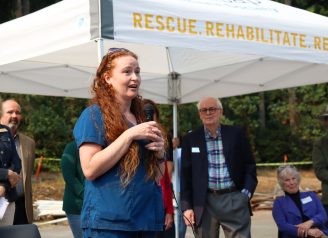 Red-haired woman speaks to a crowd with individuals gathered behind her, under a pop-up tent.